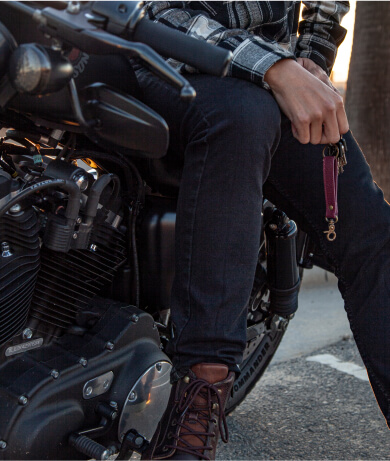 A person in a plaid shirt and jeans sits astride a black motorcycle, hand resting on the knee, on an urban road.