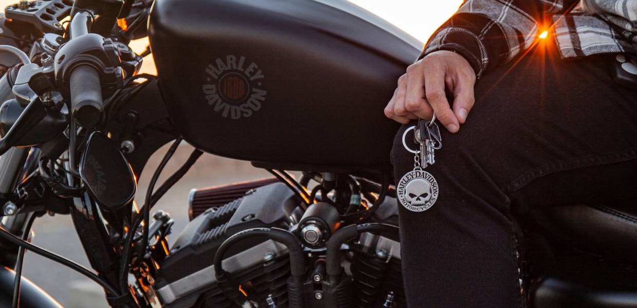 A person is holding a keyring with the Harley-Davidson logo, near a motorcycle with matching branding, during sunset.