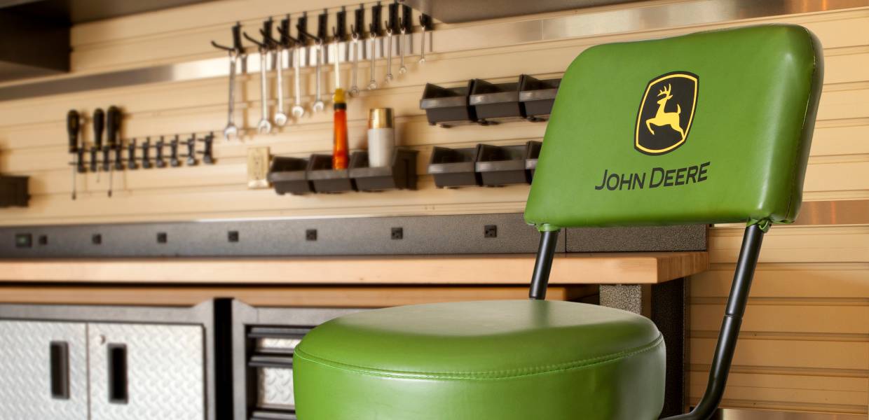 A green chair with "John Deere" logo on the backrest sits in a tidy workshop with tool racks and cabinets.