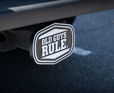 A black and white badge stating "OLD GUYS RULE" is attached to a car's rear bumper, set against an asphalt background.