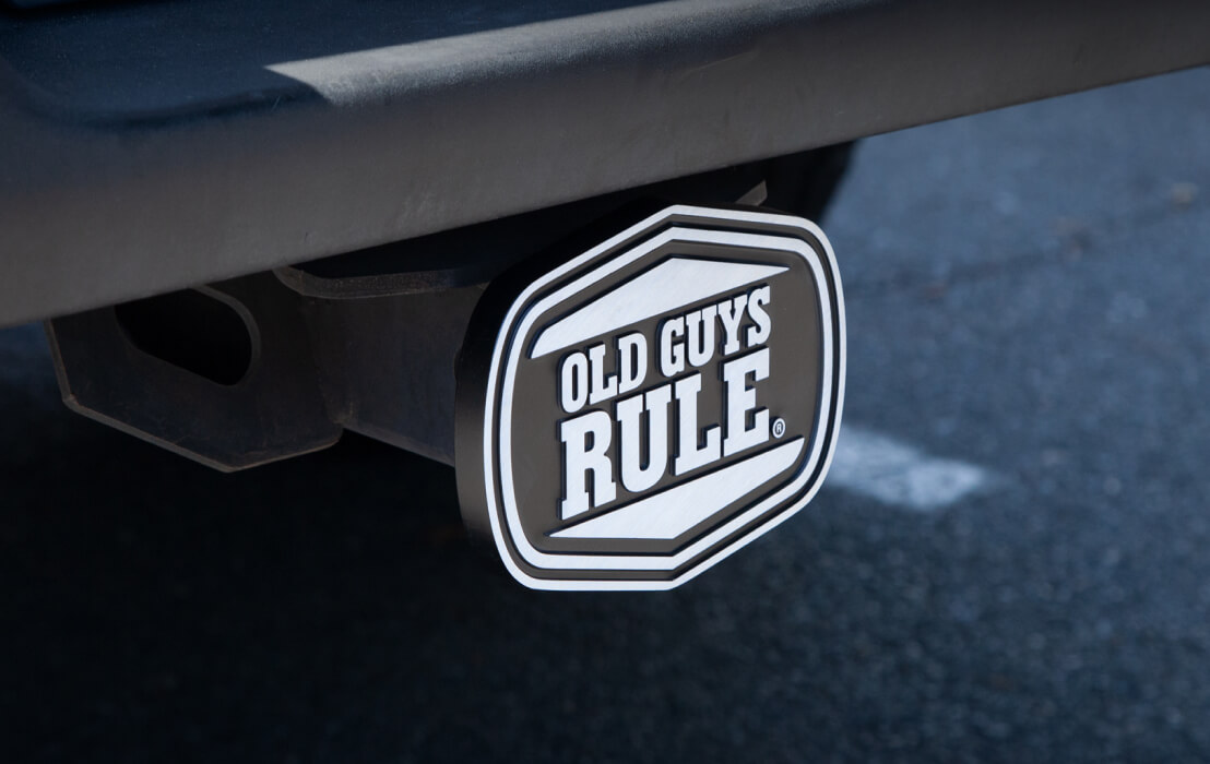 A black and white badge stating "OLD GUYS RULE" is attached to a car's rear bumper, set against an asphalt background.