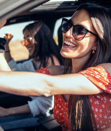 A smiling woman driving a car, another woman in the passenger seat dancing joyfully, both enjoying a sunny day on a road trip.