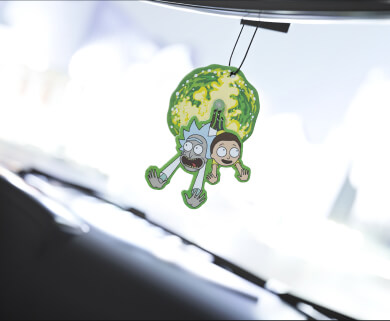 A car air freshener with a cartoon character design hangs from the rearview mirror, inside a vehicle, against a blurred snowy background visible through the windshield.