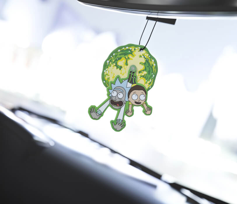 A car air freshener featuring two green, cartoonish characters resembling "Rick and Morty," dangles from a rearview mirror, with a blurred car interior in the background.